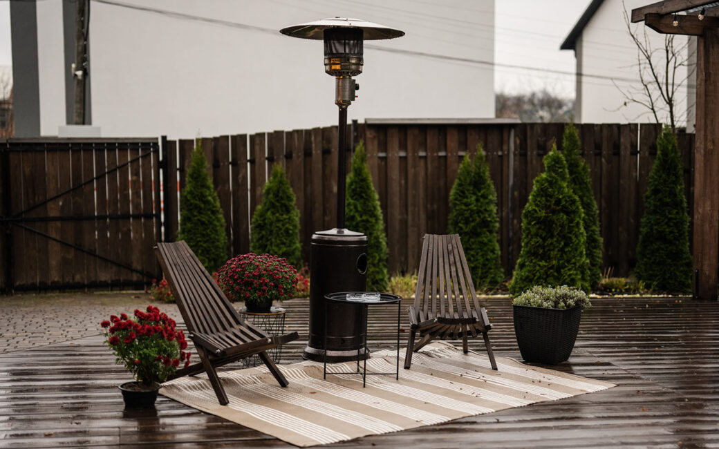 Stainless steel metal gas outdoor patio heater with wooden chairs and pot with flowers at wet terrace. Place to relax.