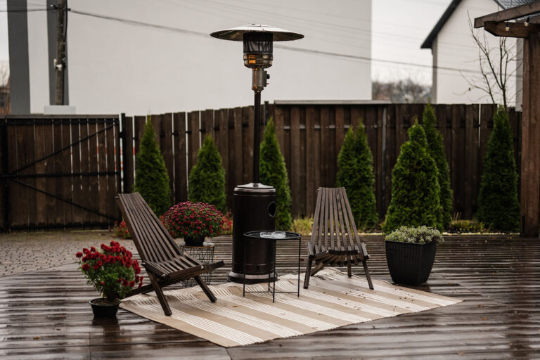 Stainless steel metal gas outdoor patio heater with wooden chairs and pot with flowers at wet terrace. Place to relax.