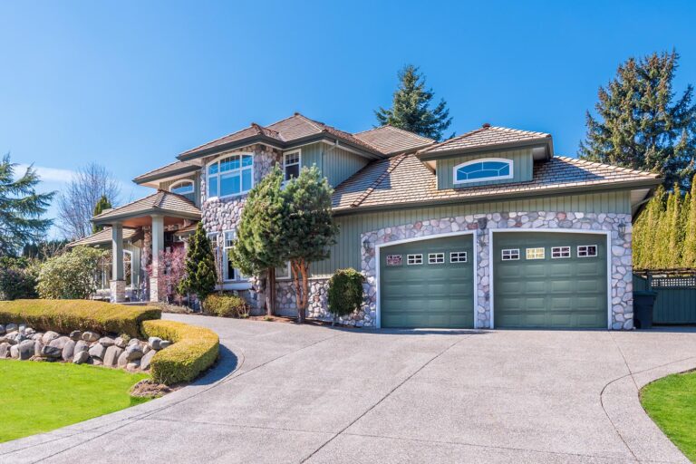 This inviting, eye-level shot presents a suburban house in daylight. The home features a two-car garage and a well-maintained lawn with trimmed bushes and various trees. A curved driveway leads up to the house, under a clear blue sky.