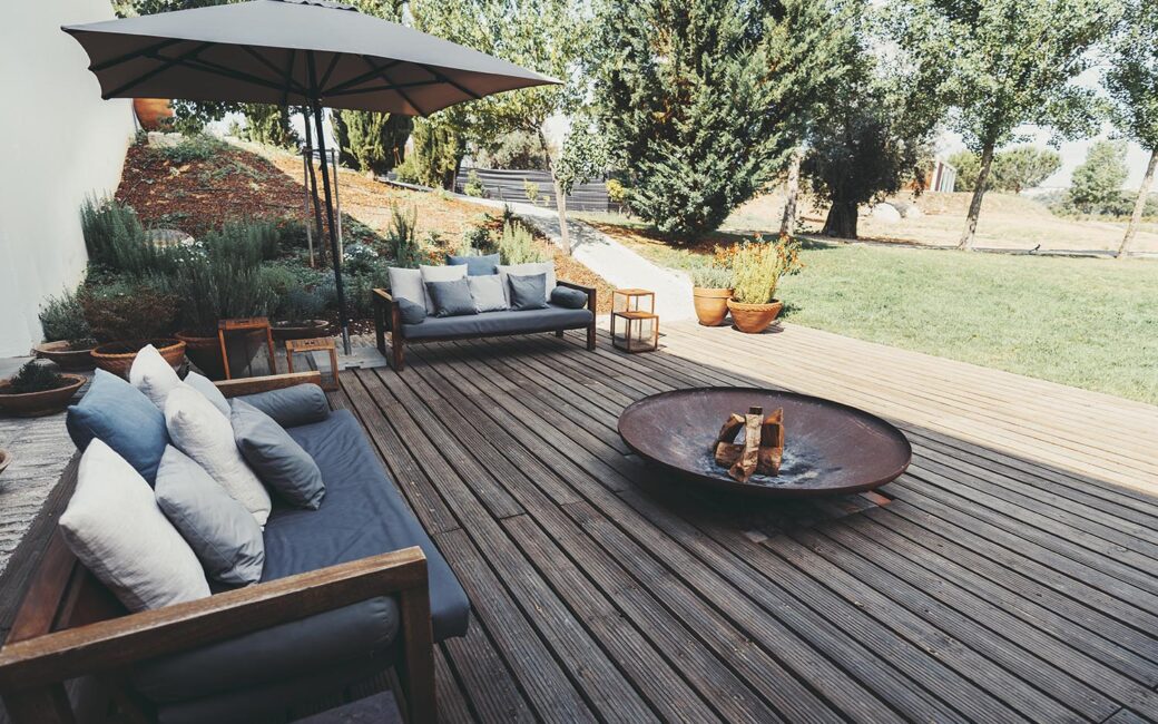 Outdoor patio area with wooden deck, cushioned sofas, and a fire pit. An umbrella provides shade, and the garden is landscaped with various plants and trees, creating a cozy ambiance