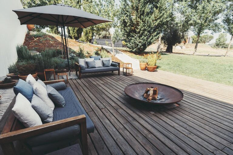 Outdoor patio area with wooden deck, cushioned sofas, and a fire pit. An umbrella provides shade, and the garden is landscaped with various plants and trees, creating a cozy ambiance