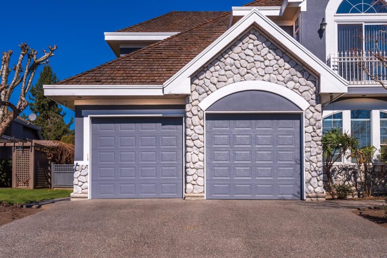 A view of a suburban house on a sunny day. The home features two garage doors, stone accents, and a brown shingled roof.
