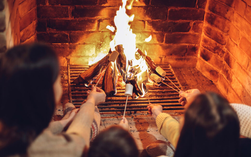 Happy young family parents with two small kids gathering around brick fireplace in cozy and warm country house during Christmas holidays in winter. Father, mother ad children sitting by fire at home