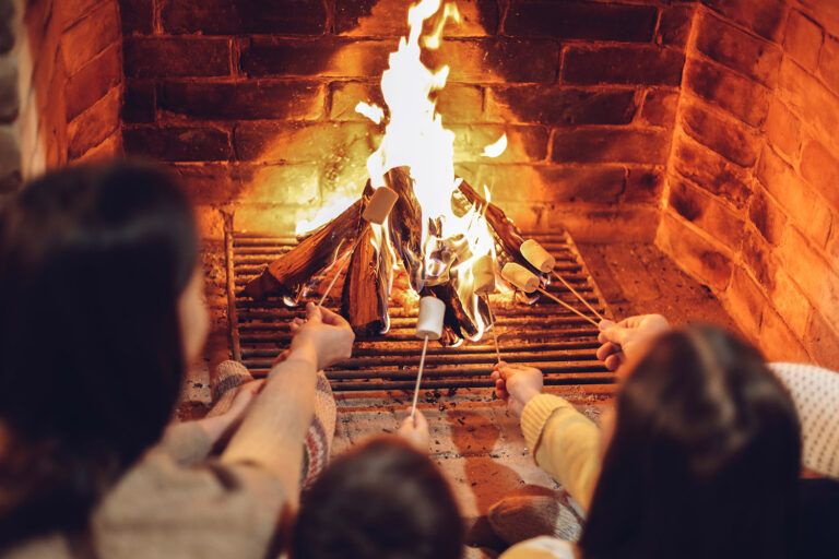 Happy young family parents with two small kids gathering around brick fireplace in cozy and warm country house during Christmas holidays in winter. Father, mother ad children sitting by fire at home