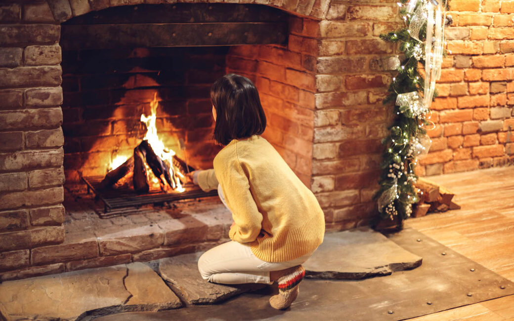 Little girl toasting marshmallow on chopstick over fireplace decorated with traditional xmas ornaments, rear view. Child near brick fireside in cozy house during winter holidays. Christmas coziness