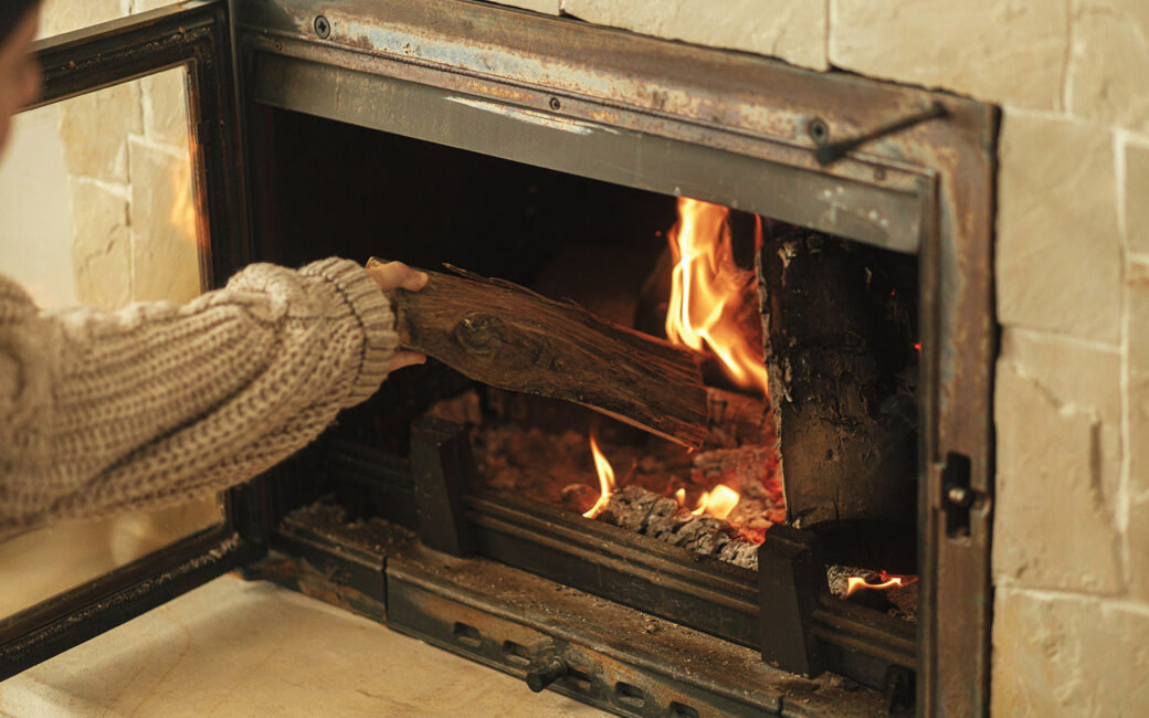 Woman putting firewoods into burning fireplace in rustic room in farmhouse. Heating house in winter with wood burning stove. Fireplace heating alternative to gas and electricity
